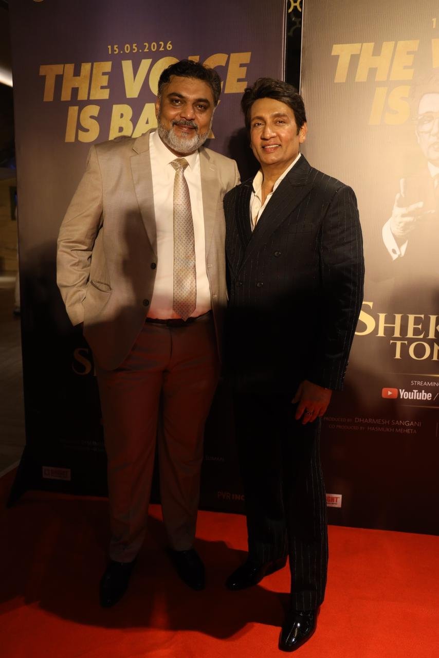 Two men in suits posing on a red carpet at a promotional backdrop that reads 'THE VOICE IS BACK'.